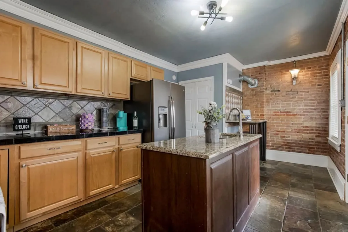 The photo features a granite kitchen island, stainless steel appliances, and an exposed brick wall. The inviting atmosphere suggests shared activities like cooking or enjoying a meal together.