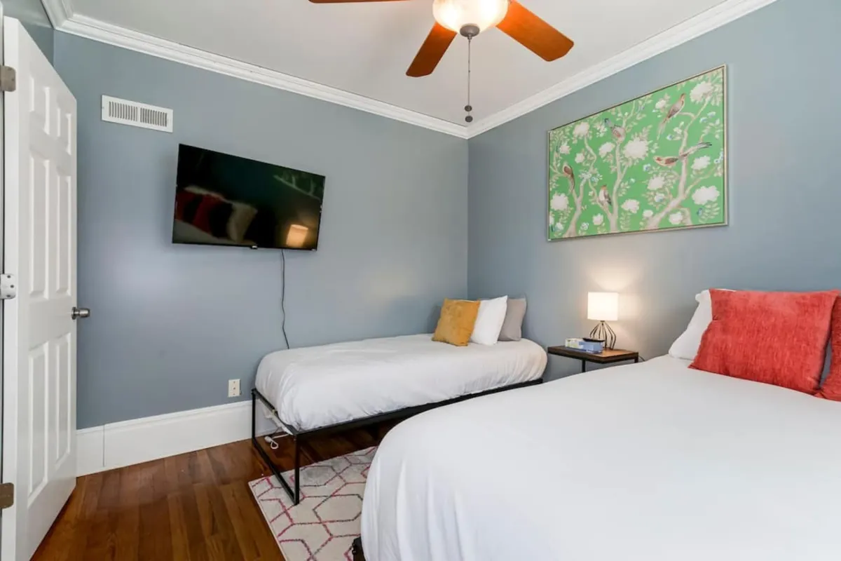 A serene bedroom featuring two beds, wall art, a ceiling fan, and a TV. The space is calm and uncluttered, with soft morning light and amenities for both relaxation and entertainment, making it suitable for families or multiple guests.