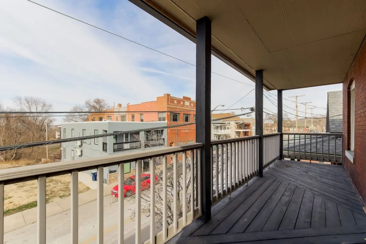 An exterior balcony view with a sturdy railing overlooks the street and surrounding buildings. The space is set up for casual enjoyment, such as sipping morning coffee or people-watching, suggesting an independent, self-service lifestyle.