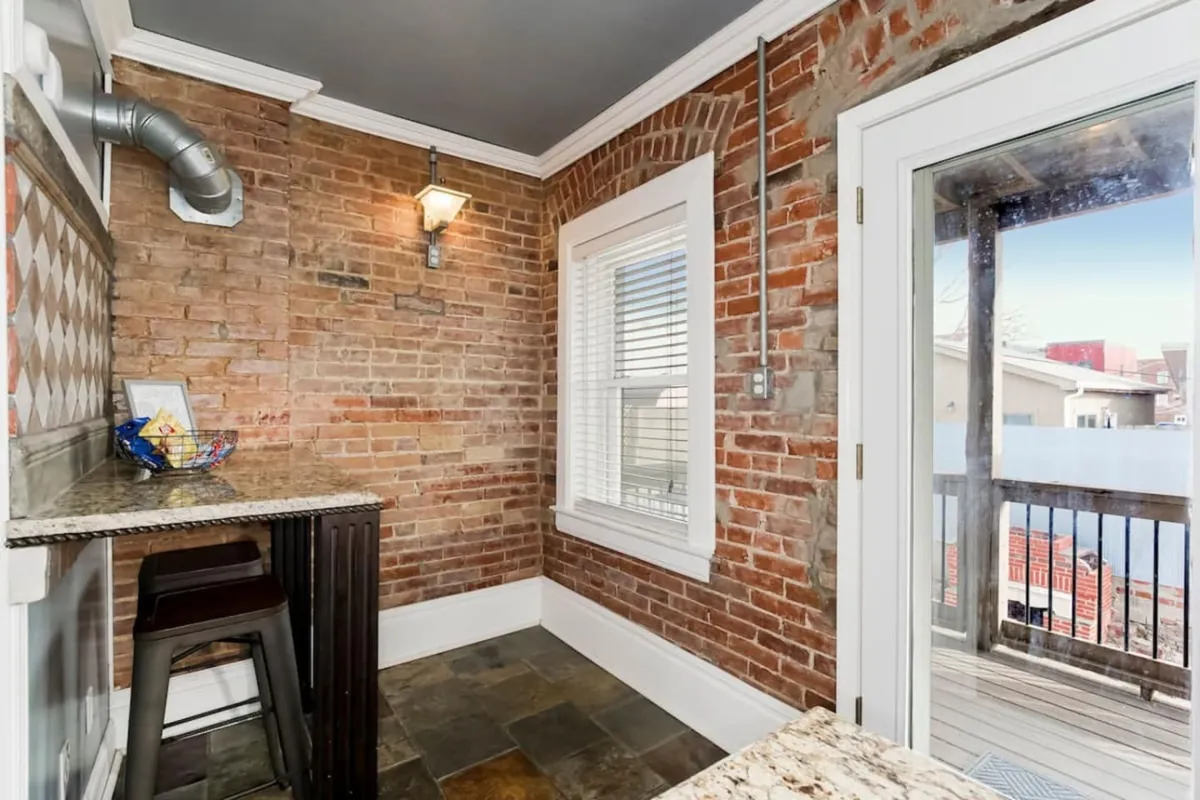 A bright and airy kitchen/dining area with exposed brick, a granite countertop, bar stools, and abundant natural light from a large window. A door leads to a balcony, enhancing the sense of openness and connection.