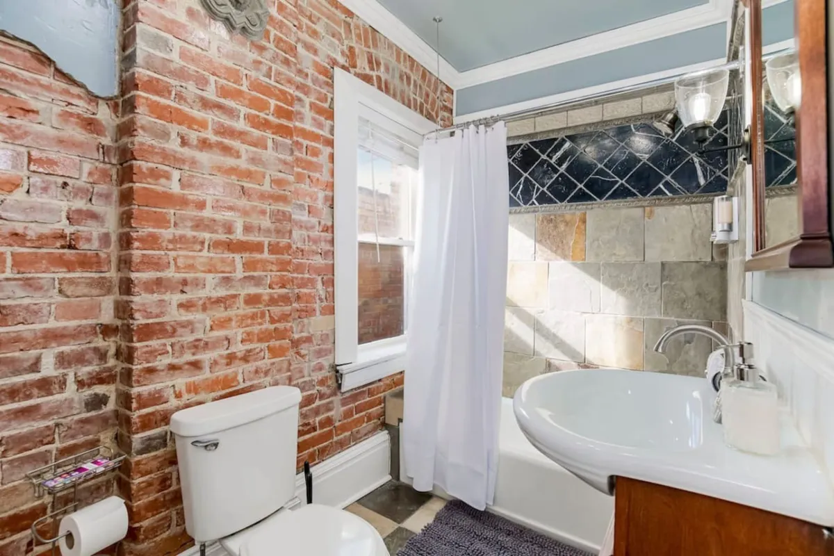 A charming bathroom scene featuring an exposed brick wall, white shower curtain, modern sink, and slate tiles, with sunlight filtering in to create a cozy morning atmosphere.