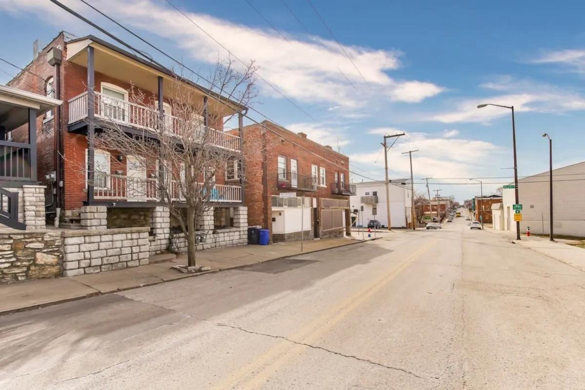 The image captures vibrant red brick buildings with balconies, a tree-lined street, and signs of community life such as people walking and shopping. The lively outdoor setting hints at the dynamic neighborhood guests can explore.