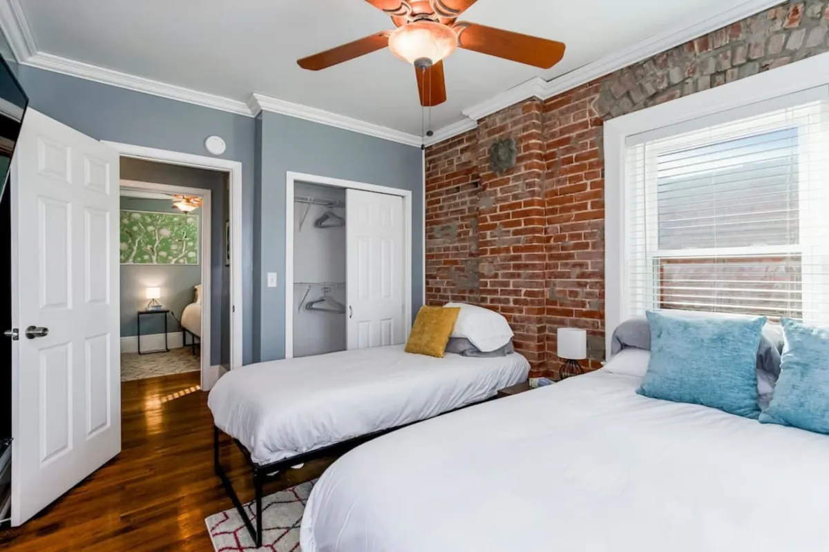 A welcoming bedroom with two beds set against an exposed brick wall, featuring soft lighting, a ceiling fan, and a cozy reading nook by the window. Sunlight streams in, creating a warm, restful atmosphere ideal for unwinding after a busy day.