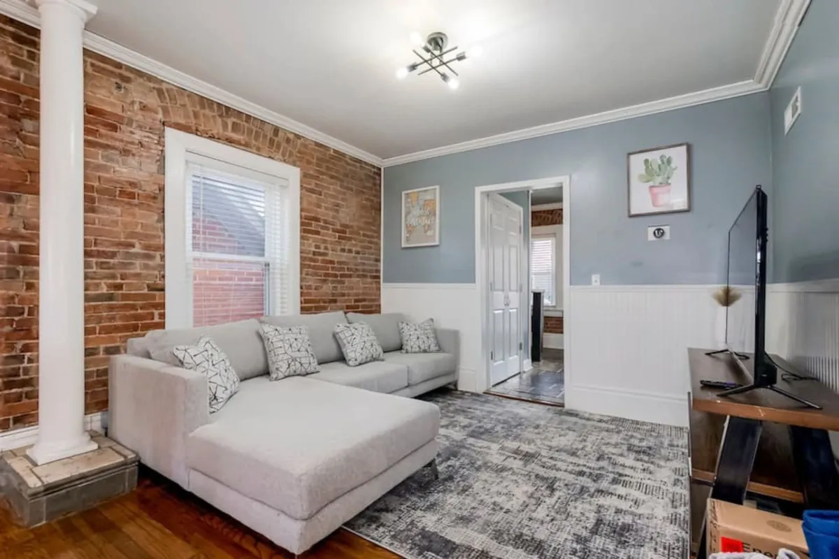 A cozy living room with a plush sofa facing a flat-screen television, exposed brick wall backdrop, and decorative artwork. The setup encourages socializing, watching TV, and relaxing in an intimate environment.