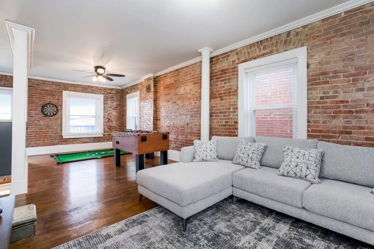 A spacious open-plan living area featuring a comfy sectional sofa, foosball table, large windows letting in natural sunlight, and exposed brick walls. The room is set up for relaxation, socializing, and group activities, with ample seating and an inviting, casual atmosphere.