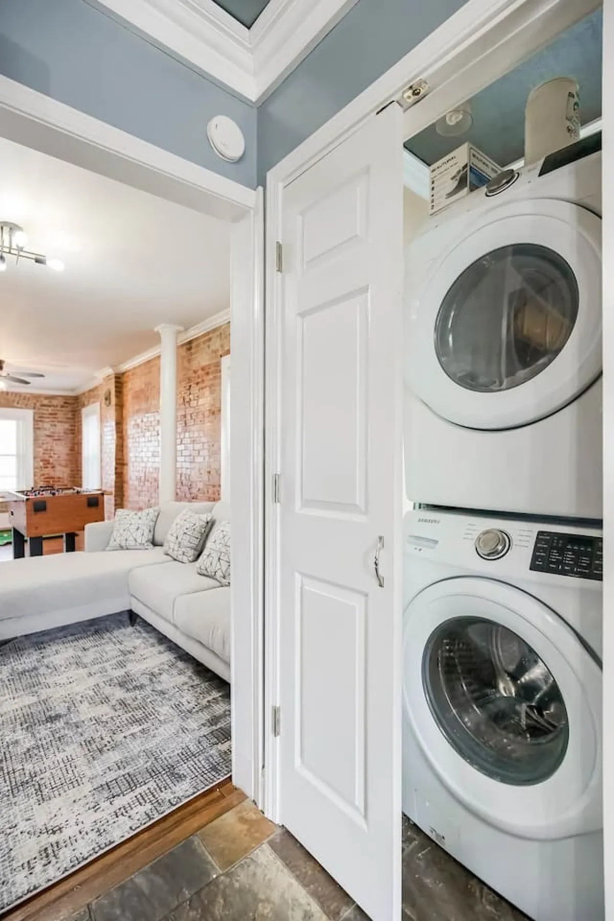A casual, comfortable interior scene featuring a washer and dryer set next to a couch and foosball table, providing a glimpse of a functional laundry area integrated into a relaxed living space.