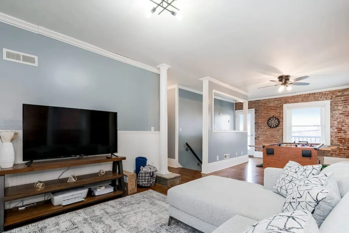 The image showcases a stylish, sunlit living room with a plush sofa facing a large television, set against a striking exposed brick wall. A modern coffee table sits in the center, with a window allowing natural light to fill the space, creating a welcoming atmosphere perfect for families to gather, watch movies, or play games together.